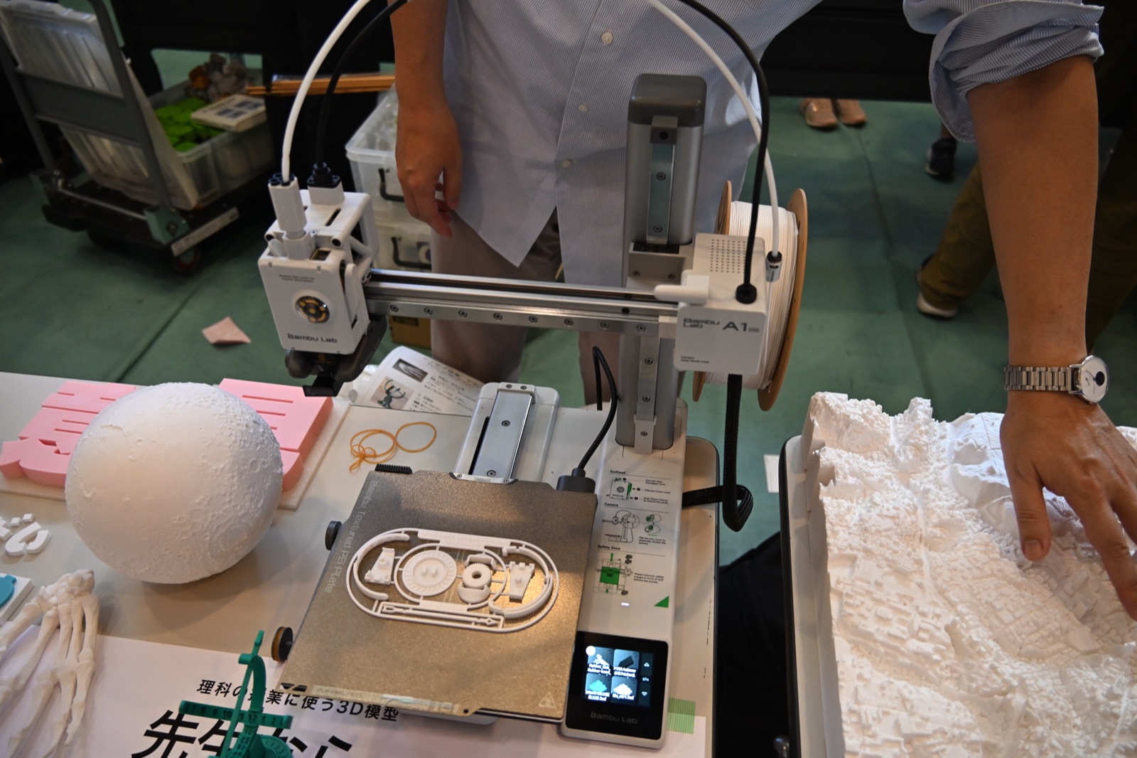 栃木の会場の机。動いている3Dプリンターのそばに月・骨・立体地形図が並び、手が立体地形図に触れている